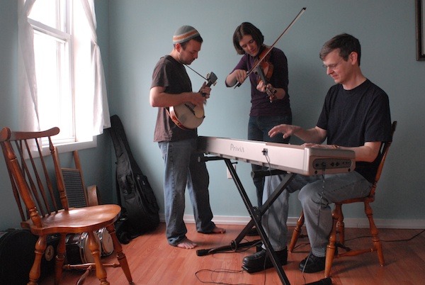 Three musicians from Contranella playing in a sunny room, Megan on fiddle, John on Piano, and Charley on mandolin